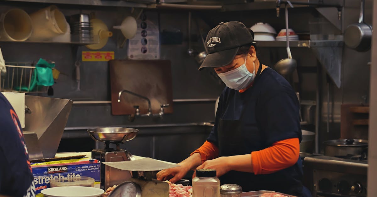 A woman in a kitchen wears a face mask while cutting meat, showcasing food preparation safety.